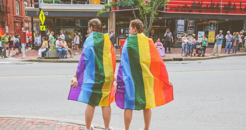 zwei Frauen eigehüllt in Regenbogenfahnen stehen an einer belebten Kreuzung