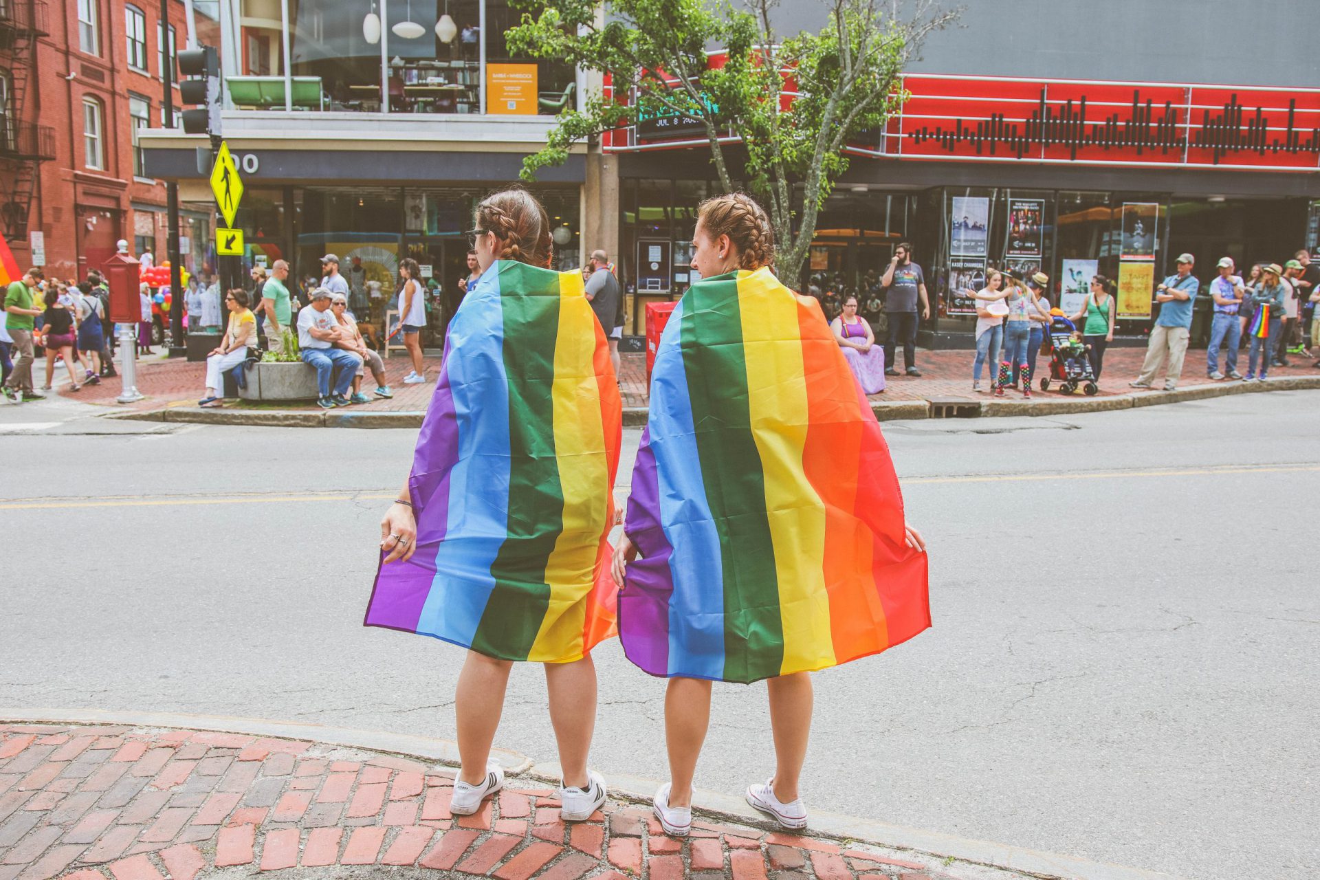 zwei Frauen eigehüllt in Regenbogenfahnen stehen an einer belebten Kreuzung
