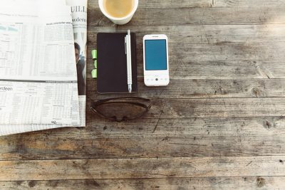 Blick auf einen rustikalen Tisch mit Zeitung, Notizbuch, Mobiltelefon und Sonnenbrille und einer Tasse Kaffee.