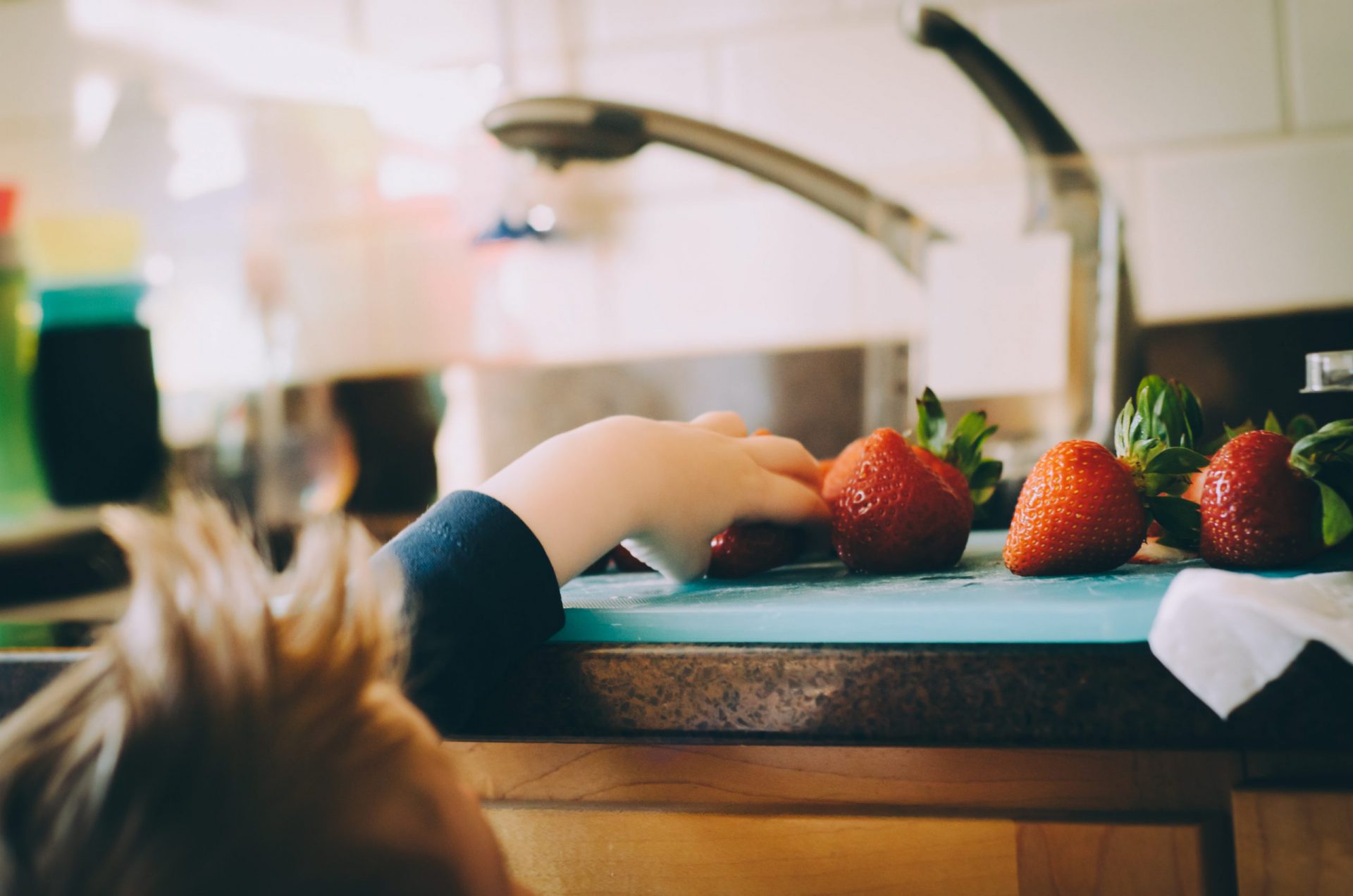 Ein Kind greift nach Erdbeeren, die neben dem Spülbescken auf einer Küchenanrichte liegen
