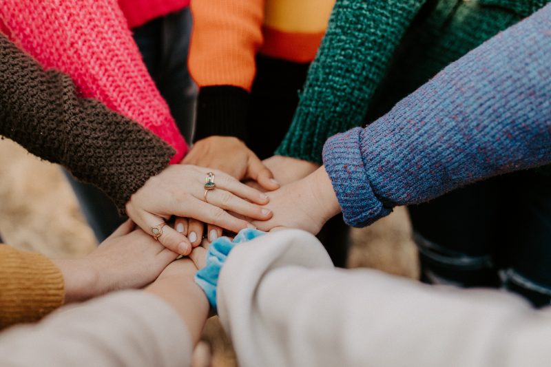 Acht Menschen in bunten Pullovern legen die Hände in der Mitte des Bildes aufeinander