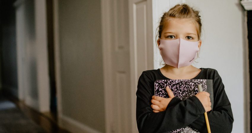 Mädchen mit rosa Mundschutz lehnt an Tür und drückt Buch und Bleistift an sich