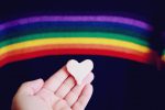 Regenbogenflagge im Hintergrund, im Vordergrund eine Hand mit Herz aus Marzipan