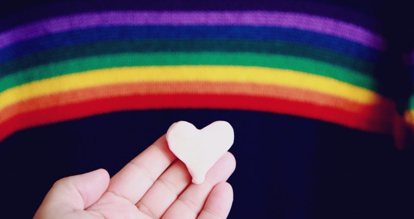Regenbogenflagge im Hintergrund, im Vordergrund eine Hand mit Herz aus Marzipan