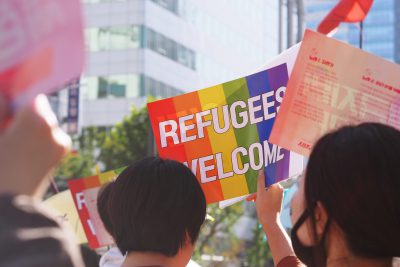 Junge Frau mit schwarzen Haaren hält Regenbogen-Plakat "Refugees welcome" hoch