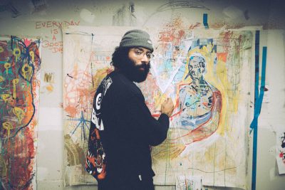 Künstler mit Vollbart, Mütze, schwarzen Pullover vor einer Leinwand, der über seine Schulter in die Kamera blickt