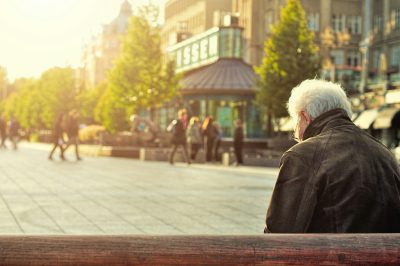 Mann mit grauen Haaren sitzt alleine auf einer Bank an einem großen Platz