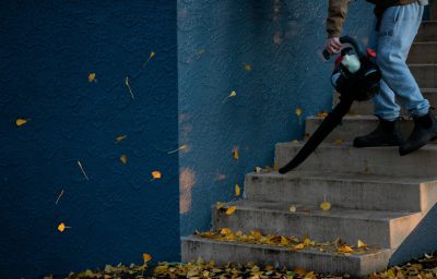 Mann in Jeans (dessen Beine man nur sieht) entfernt mit einem Laubbläser Blätter von einer Treppe