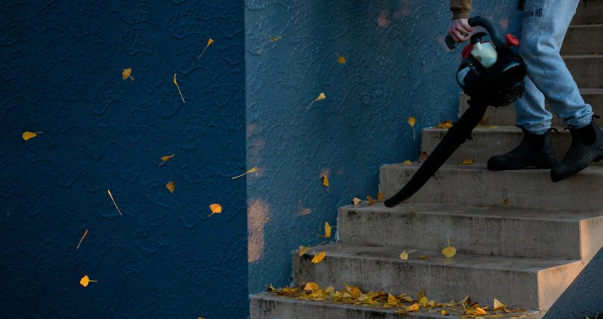 Mann in Jeans (dessen Beine man nur sieht) entfernt mit einem Laubbläser Blätter von einer Treppe